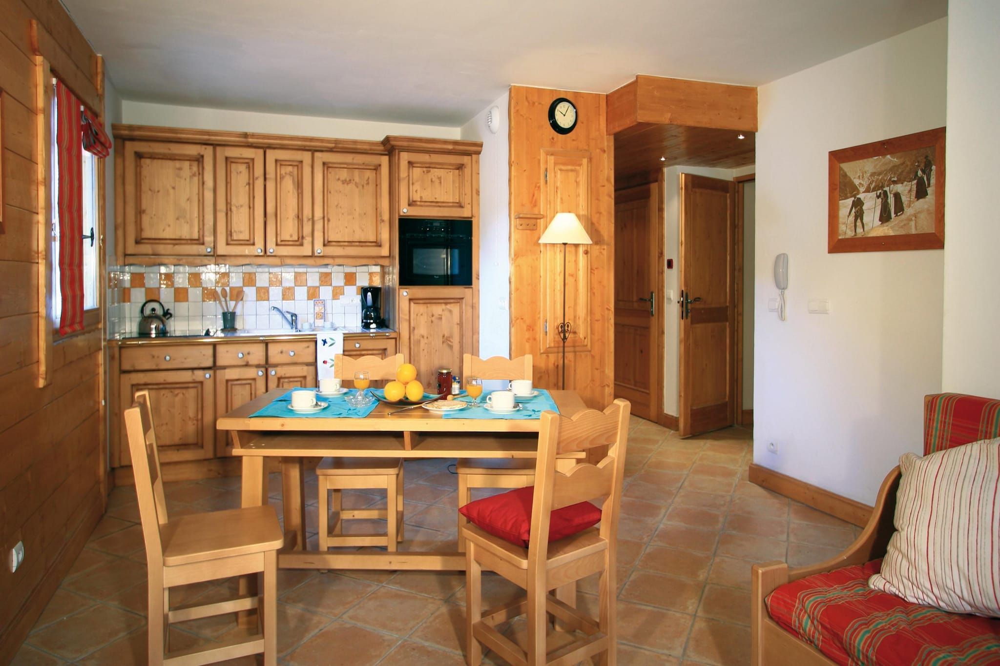 A wooden kitchen with a dining table set for two, a clock on the wall, and a painting on the wall.