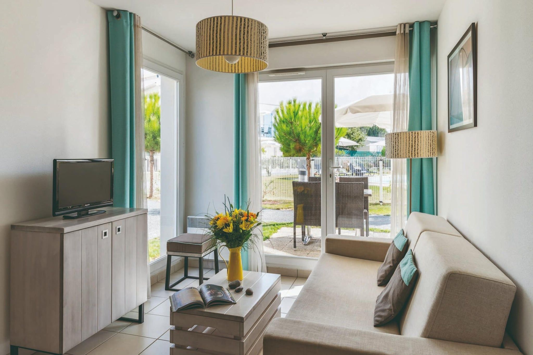 A modern living room with a sofa, a TV on a cabinet, a coffee table with a vase of flowers, and a view of an outdoor area with a fence and greenery.