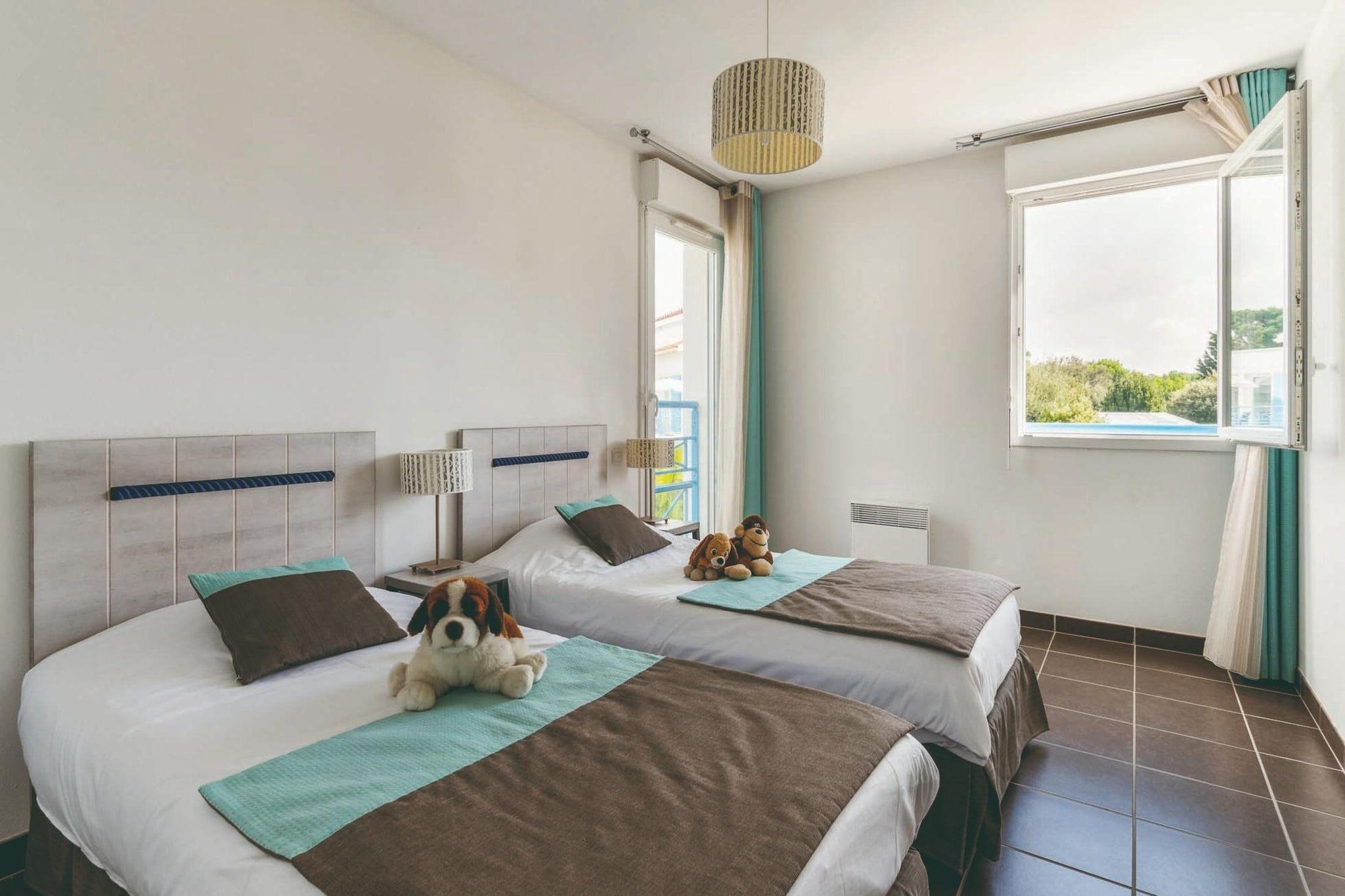 Ein Hotelzimmer mit zwei Betten, einem fensterbelaubten Fenster, einer Hängelampe und einem Stofftier auf einem Bett.