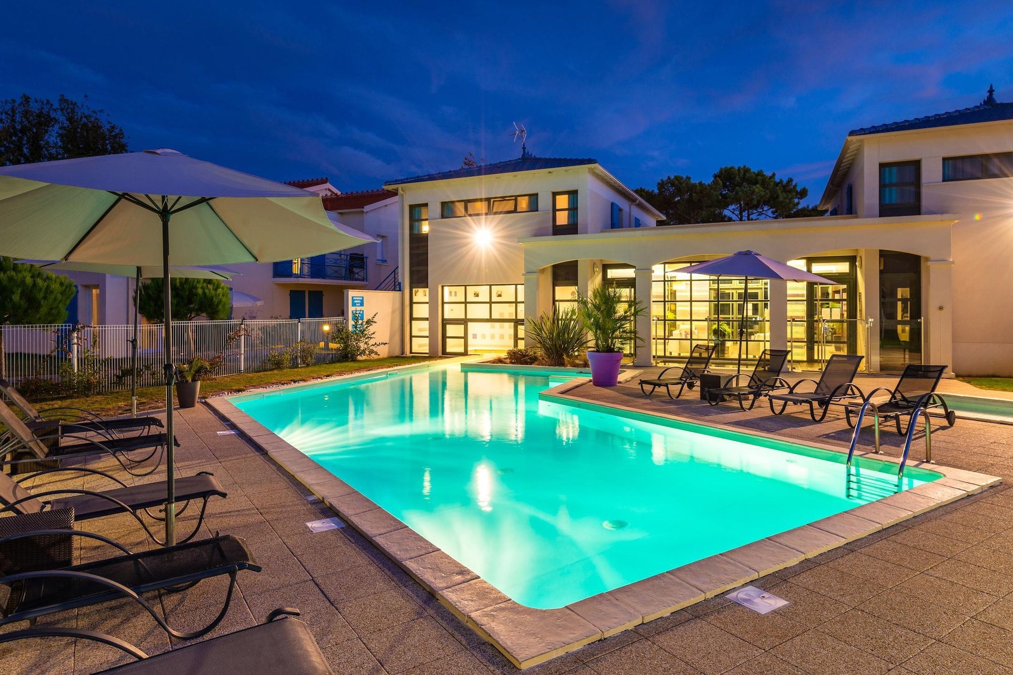 Une piscine entourée de chaises longues et de parasols, elle-même bordée d’un bâtiment doté de nombreuses fenêtres et d’une clôture.