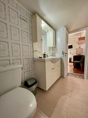 A bathroom with a toilet, sink, and mirror. There is a trash bin and a door leading to another room. - Casa Katerina studio apartment (Chania)