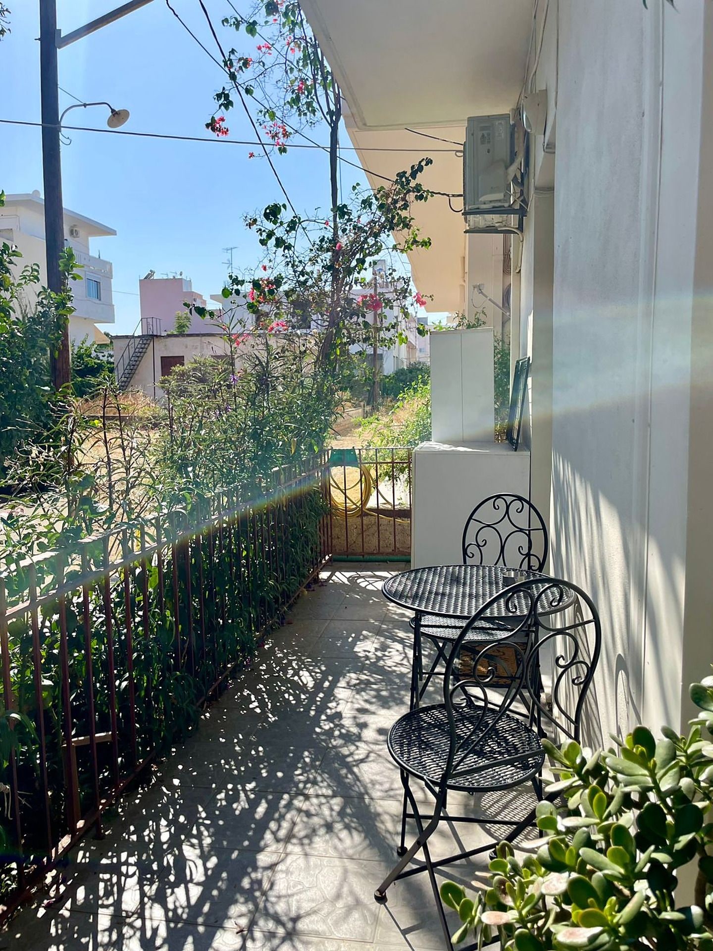 Un balcón con una mesa y sillas de metal, rodeado de follaje y vistas a la calle.