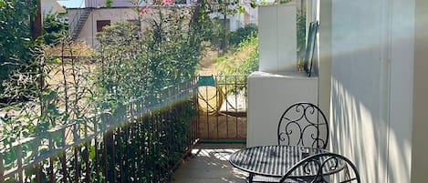 A balcony with a metal table and chairs, surrounded by greenery and a view of the street.