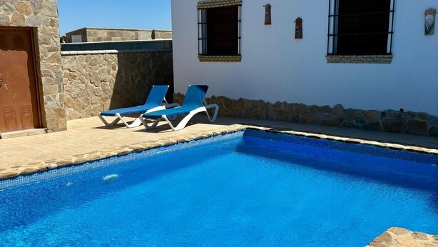 A swimming pool with two sun loungers, a stone wall, and a white building with two windows. - Casa Zerimar       . (Conil de la Frontera)