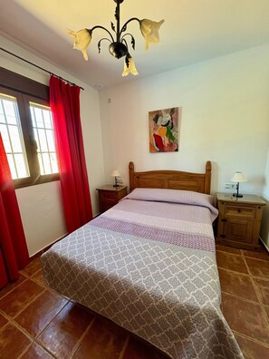 Ein Schlafzimmer mit Bett, Nachttisch, Lampe, einem Gemälde an der Wand und einem Fenster mit Vorhängen.