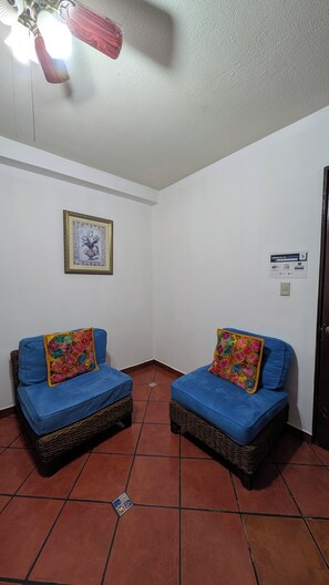 A room with two wicker sofas with blue cushions and colorful floral pillows, a ceiling fan, a framed artwork on the wall, and a door with a sign. - 101 Apartment in Villa Stahl, Colonia Escalon (San Salvador)