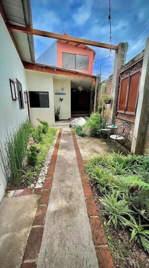 Un étroit sentier mène à un bâtiment doté d’un toit rouge, entouré de verdure et agrémenté d’un petit patio.