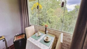 A room with a table set for two, a window with a view of trees, a small table with a vase, and a chair. - POUSADA ALL GREEN (Camanducaia)