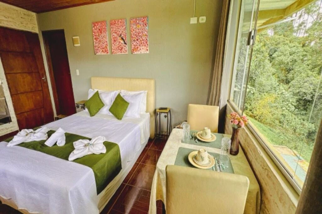 A hotel room with two beds, a small table with tea set, a chair, and a window with a view of greenery.