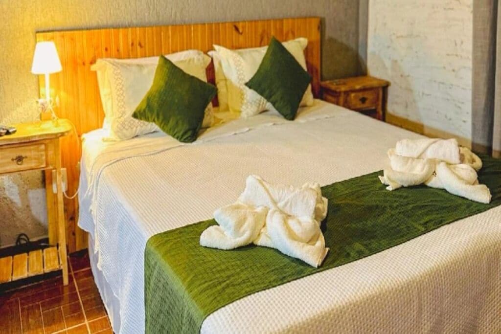 A neatly made bed with white linens and green pillows, a bedside lamp, and a small wooden nightstand with a lamp.