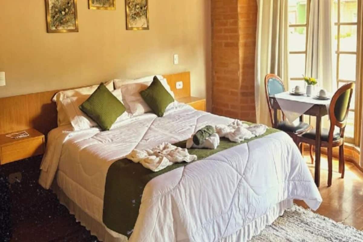 A neatly made bed with pillows, a bedside table, a chair, and a table with a vase of flowers.