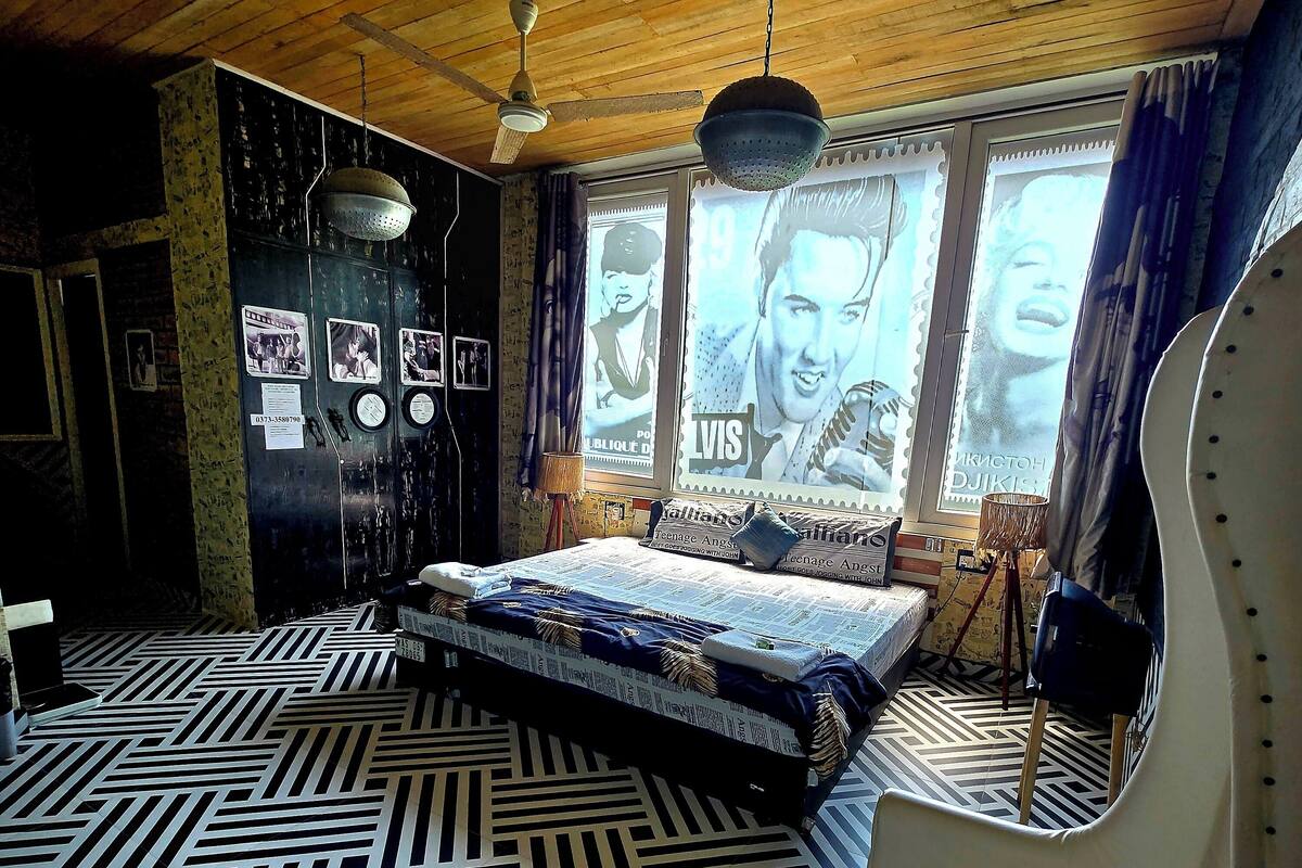 Una habitación con una cama, una silla, un ventilador de techo y carteles en la pared.