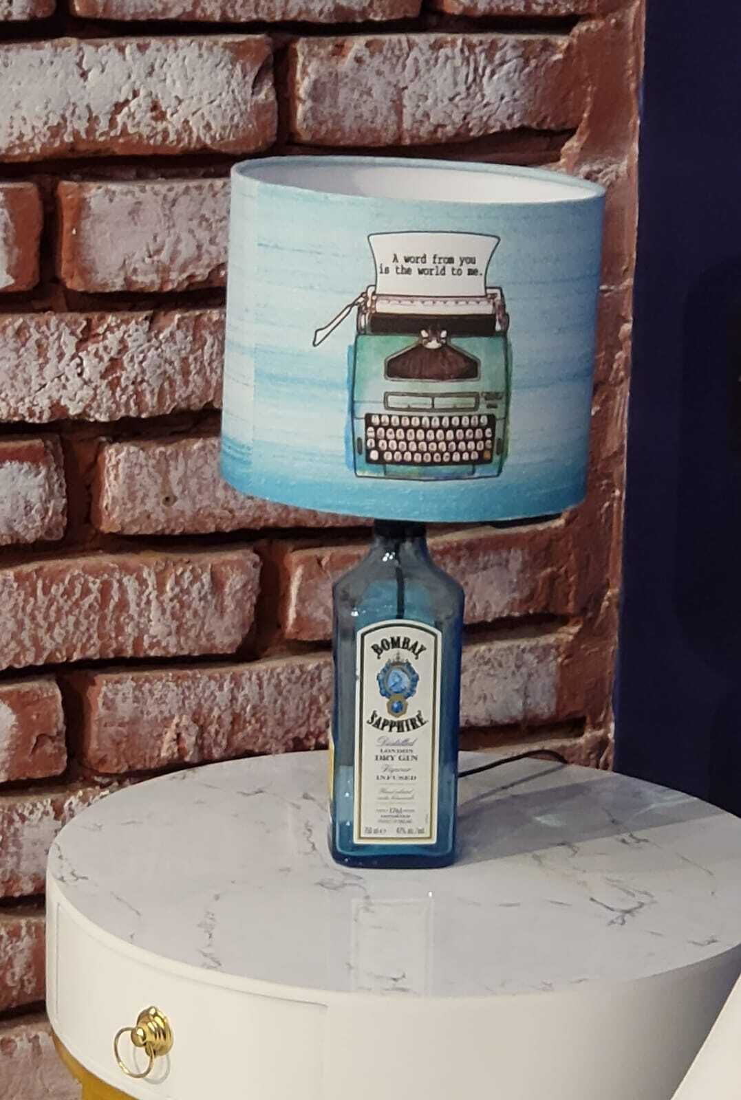 Eine Lampe mit einer Schreibmaschinen-Applikation am Schirm, eine Flasche Bombay Sapphire Gin und eine Ziegelwand im Hintergrund.