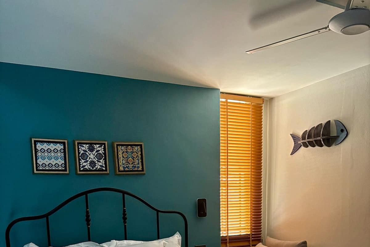 A bed with white linens, a bedside table, a wall-mounted fish decoration, and a window with blinds.