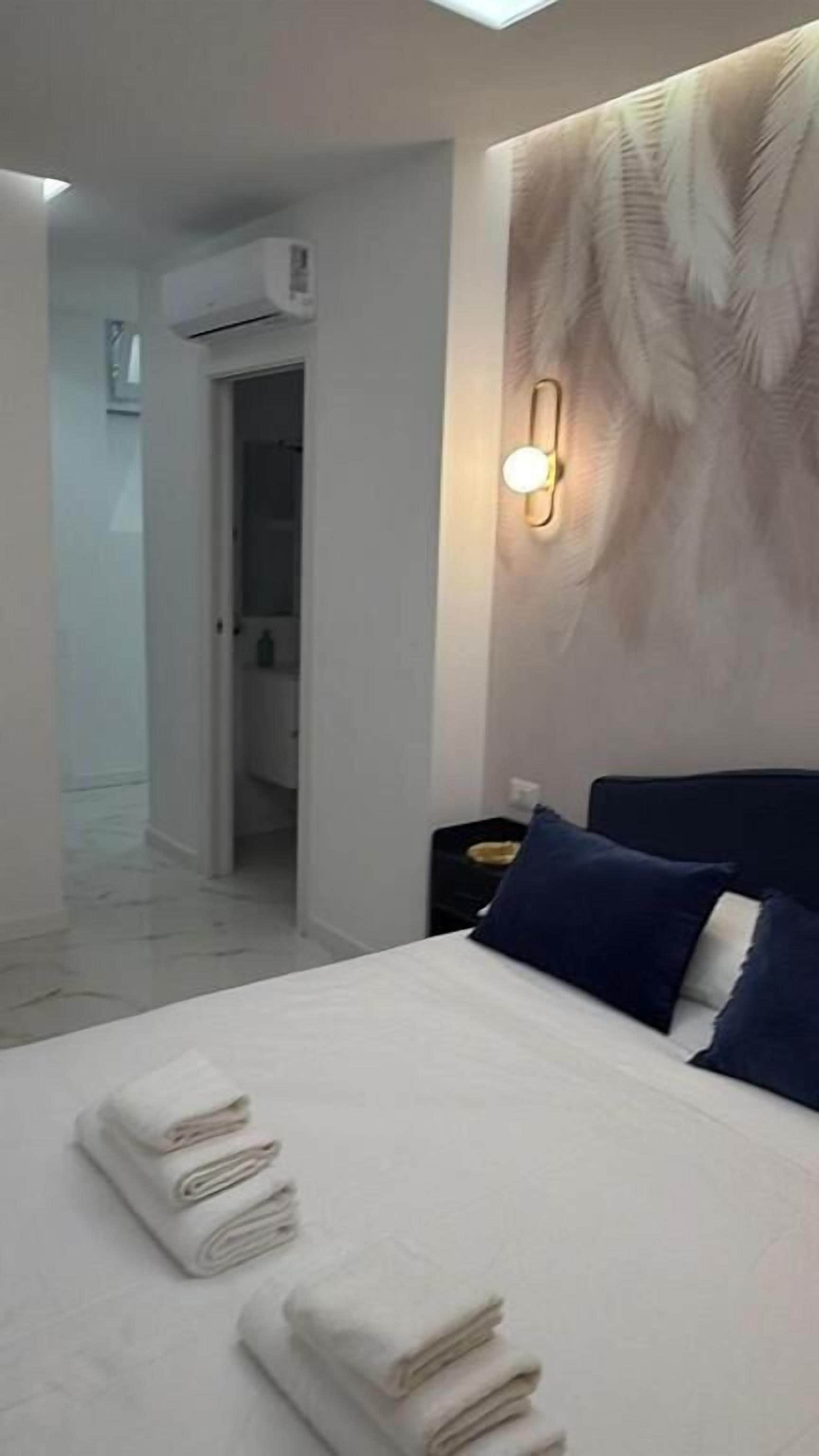 Hotellihuone, jossa on sänky, pyyhkeet, yöpöytä ja seinään kiinnitetty valaisin.