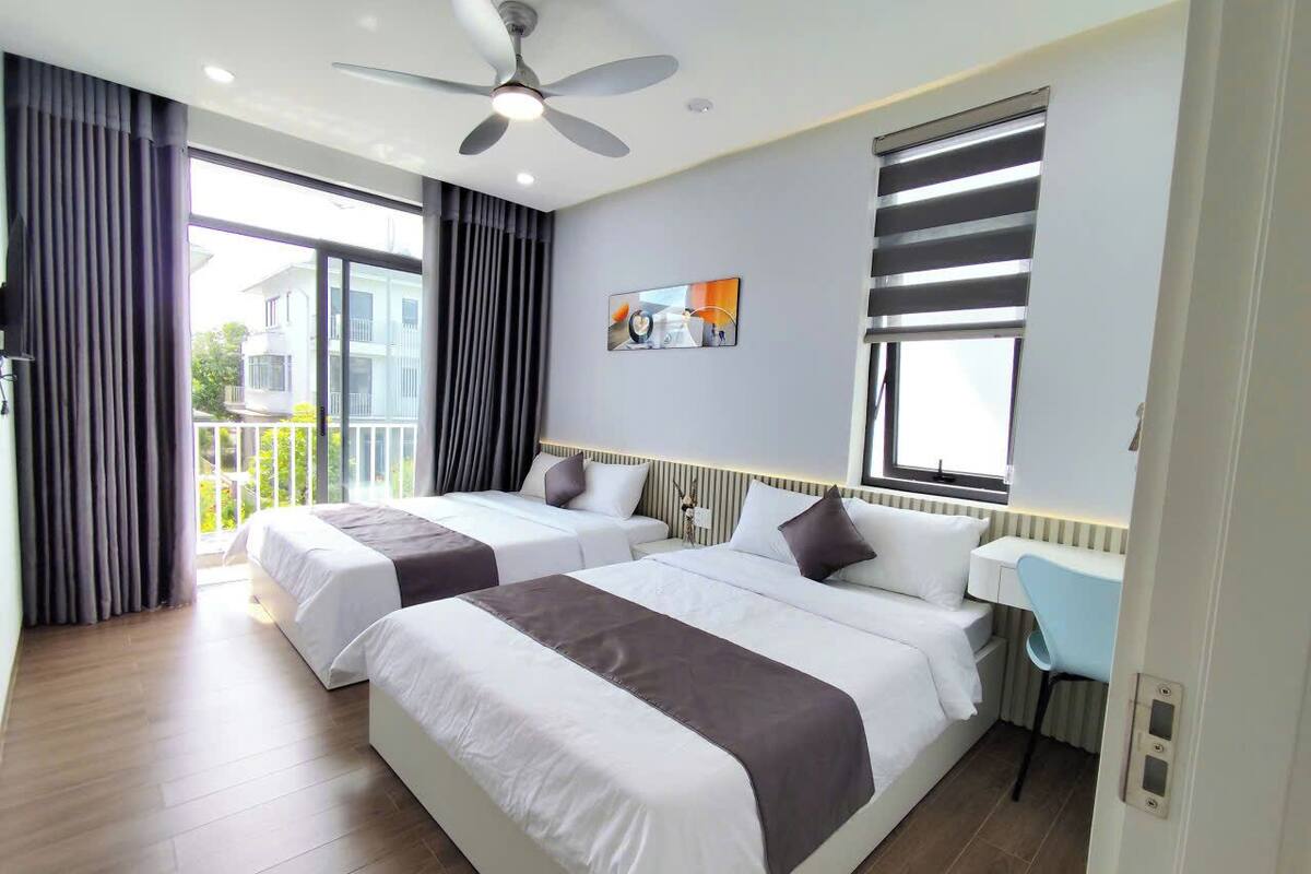 Habitación de hotel con dos camas, ventilador de techo, ventana con persianas, escritorio y vistas al exterior.