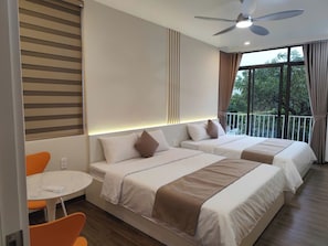 Habitación de hotel con dos camas, ventilador de techo, balcón con cortinas y una pequeña mesa con una silla.