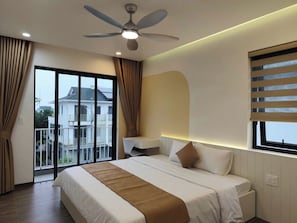 Habitación de hotel con una cama grande, un escritorio, una silla, un ventilador de techo y vistas a un edificio a través de la ventana.