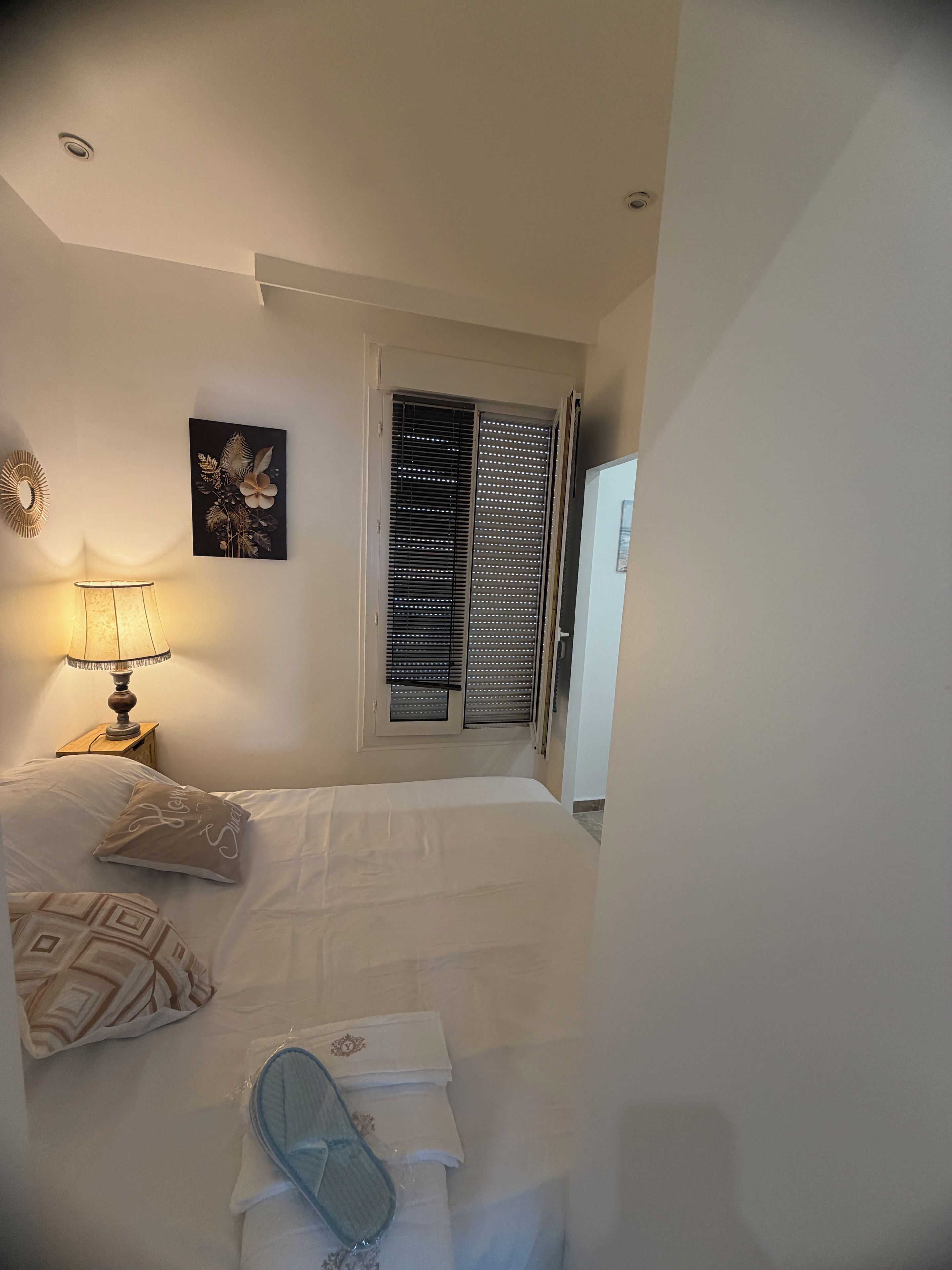 Een slaapkamer met een bed, een nachtkastje met een lamp, een raam met jaloezieën en een aan de muur hangend schilderij.
