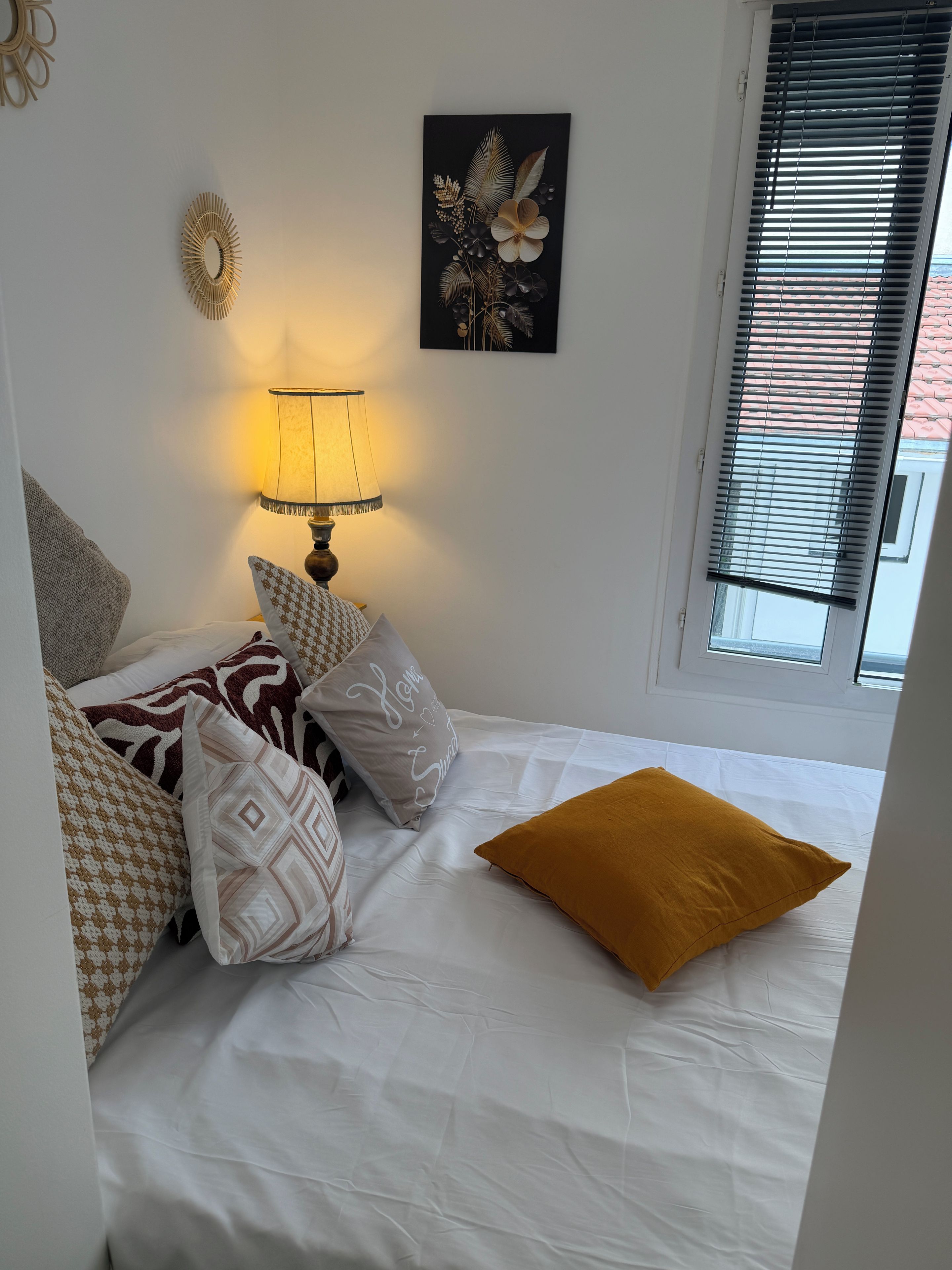 A bedroom with a bed, pillows, a bedside lamp, a wall art piece, and a window with blinds.