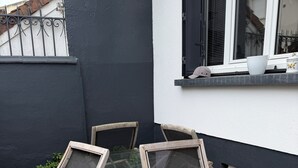 En patio med et bord og stole, et vindue med skodder og et trappetrin, der fører til en dør.