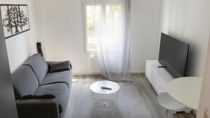 A modern living room with a grey sofa, a round coffee table, a TV on a stand, and a window with sheer curtains. - Cosy air-conditioned apartment 5 minutes from the sea (Nice)