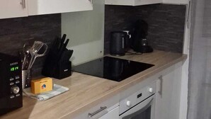 A modern kitchen with white cabinets, a black induction cooktop, a coffee maker, and a clock displaying 19:14. - Cosy air-conditioned apartment 5 minutes from the sea (Nice)
