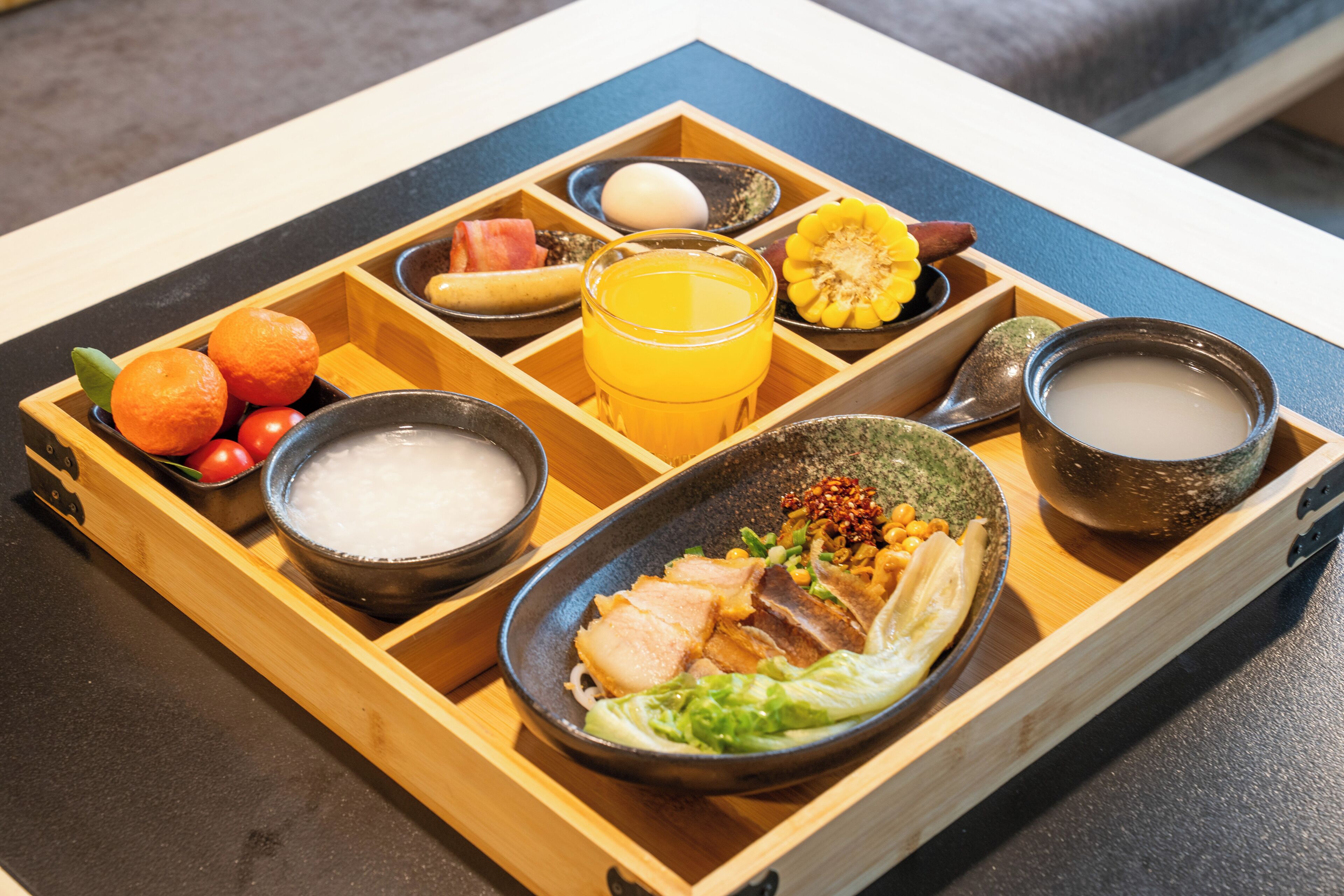 Una bandeja de madera con diversos alimentos y bebidas, entre ellos, un tazón de sopa, un plato de carne y un vaso de jugo de naranja.