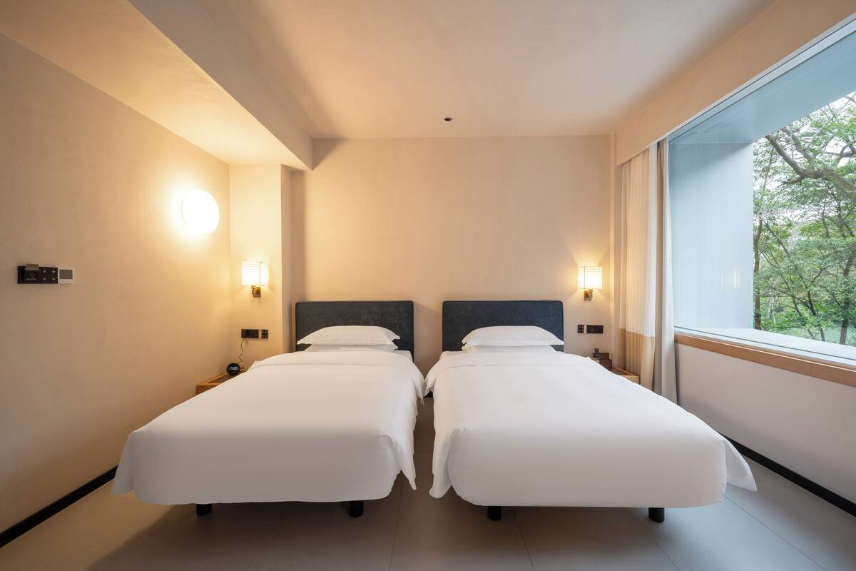 Habitación de hotel con dos camas, lámparas de noche, una ventana con vistas a árboles y una lámpara de techo.