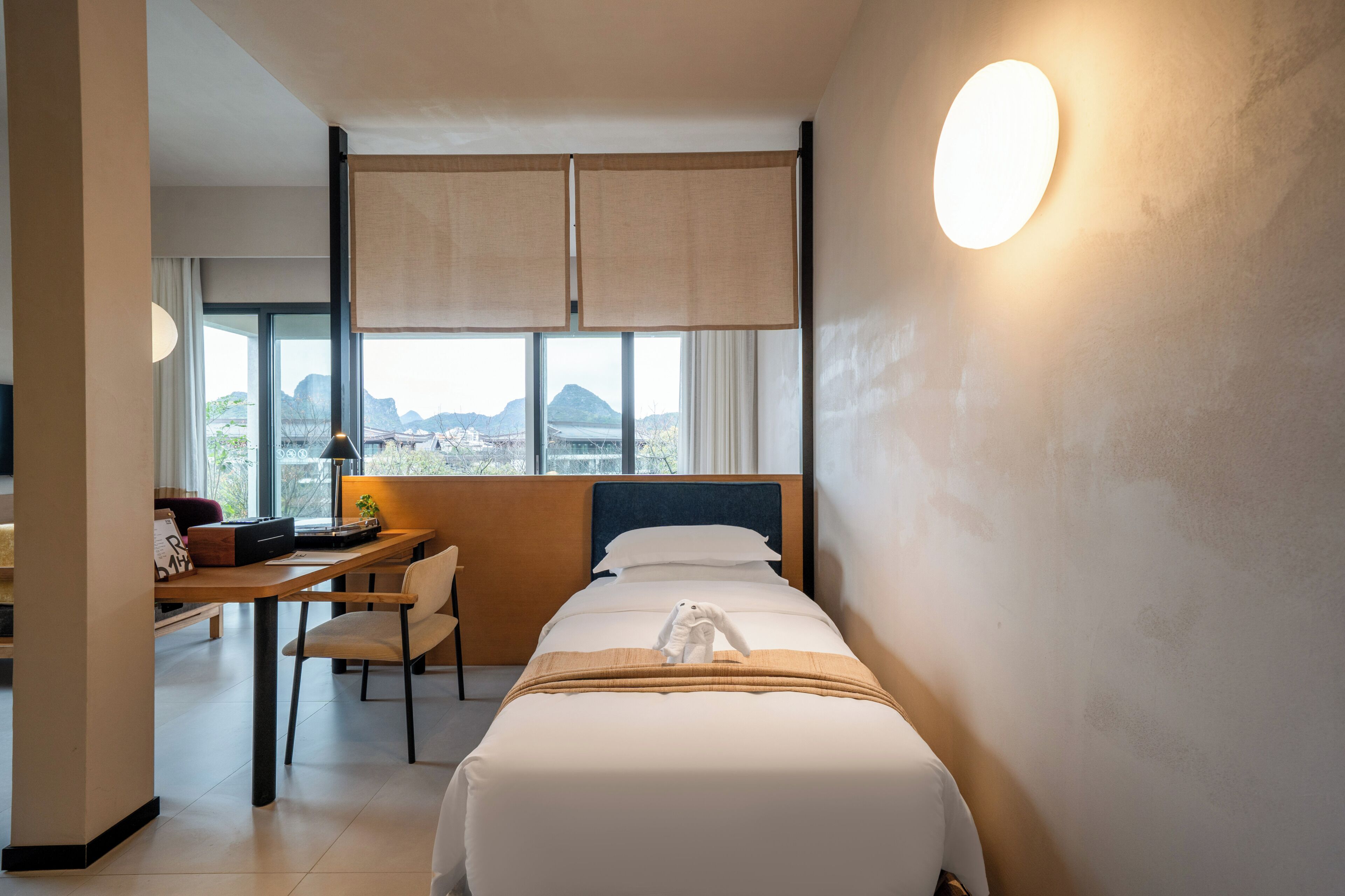 A hotel room with a bed, a desk, a chair, a lamp, and a view of mountains through the window.