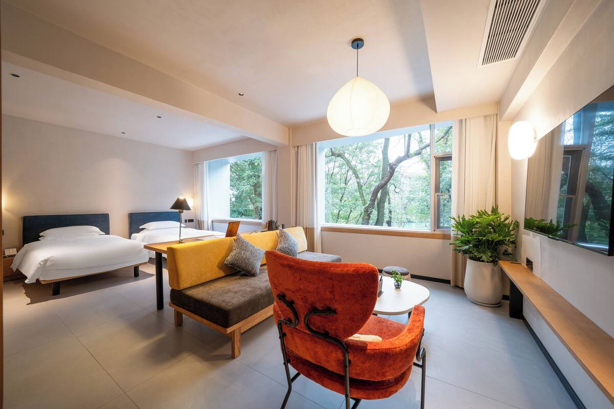Una habitación de hotel moderna con cama, sofá, mesa de comedor y un amplio ventanal con vistas a árboles.