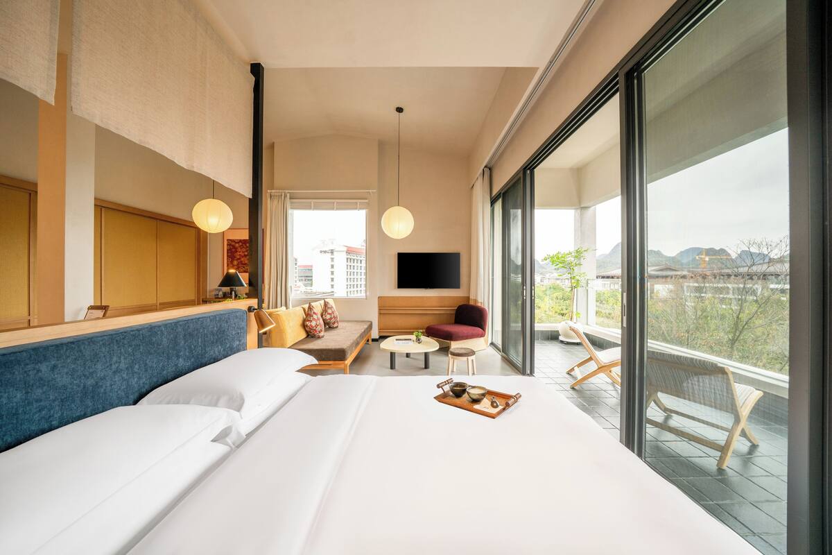 Habitación de hotel con una cama grande, un sofá, una mesita, un televisor y un balcón con vistas.