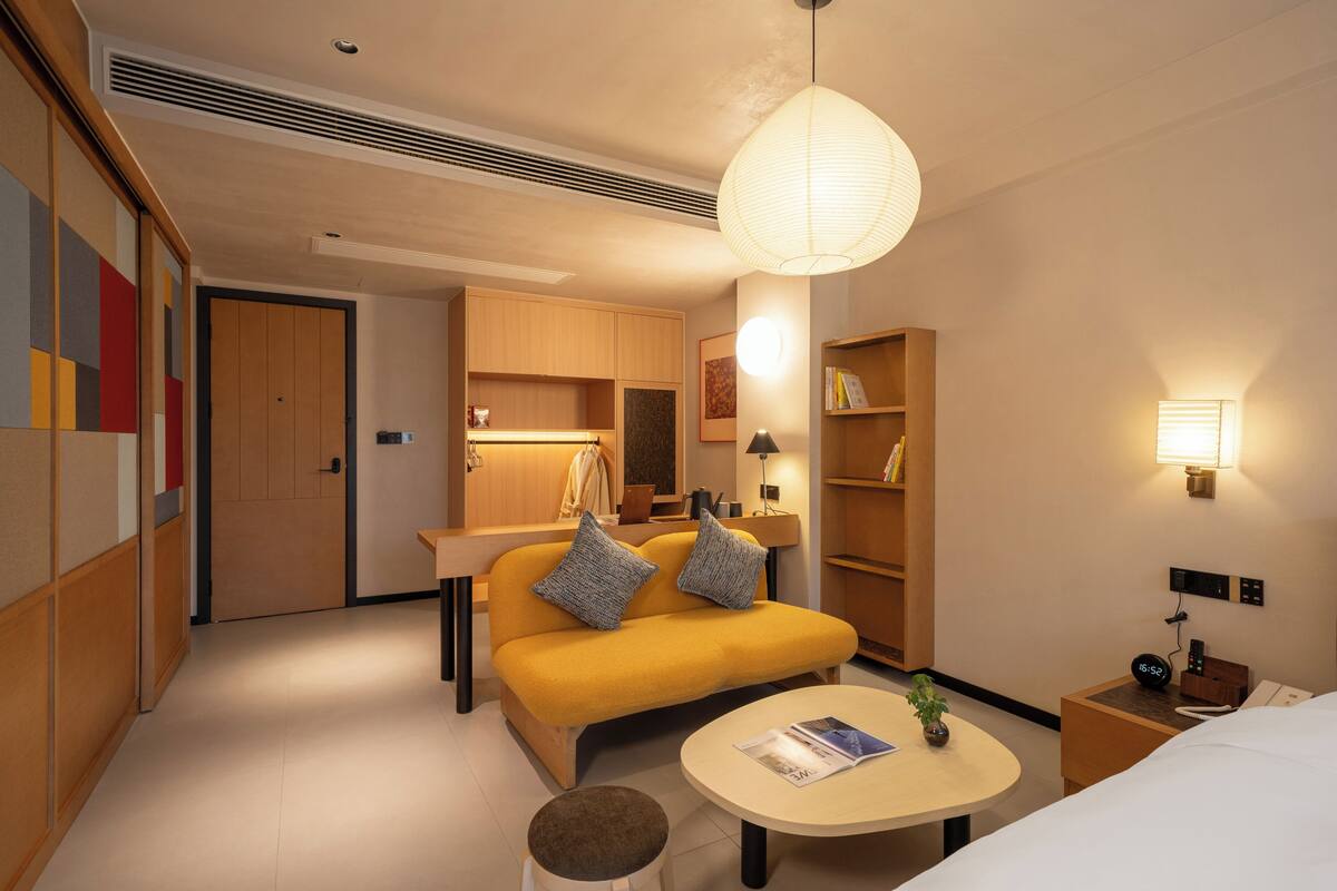 Habitación de hotel con sofá, una mesita, una mesita de noche con lámpara, estantería y armario.