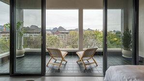 Un balcón con dos sillas de madera, una cama y vistas a montañas y edificios.