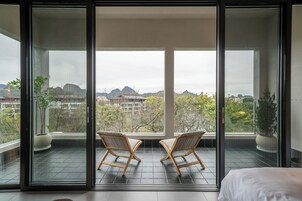 A balcony with two wooden chairs, a bed, and a view of mountains and buildings.