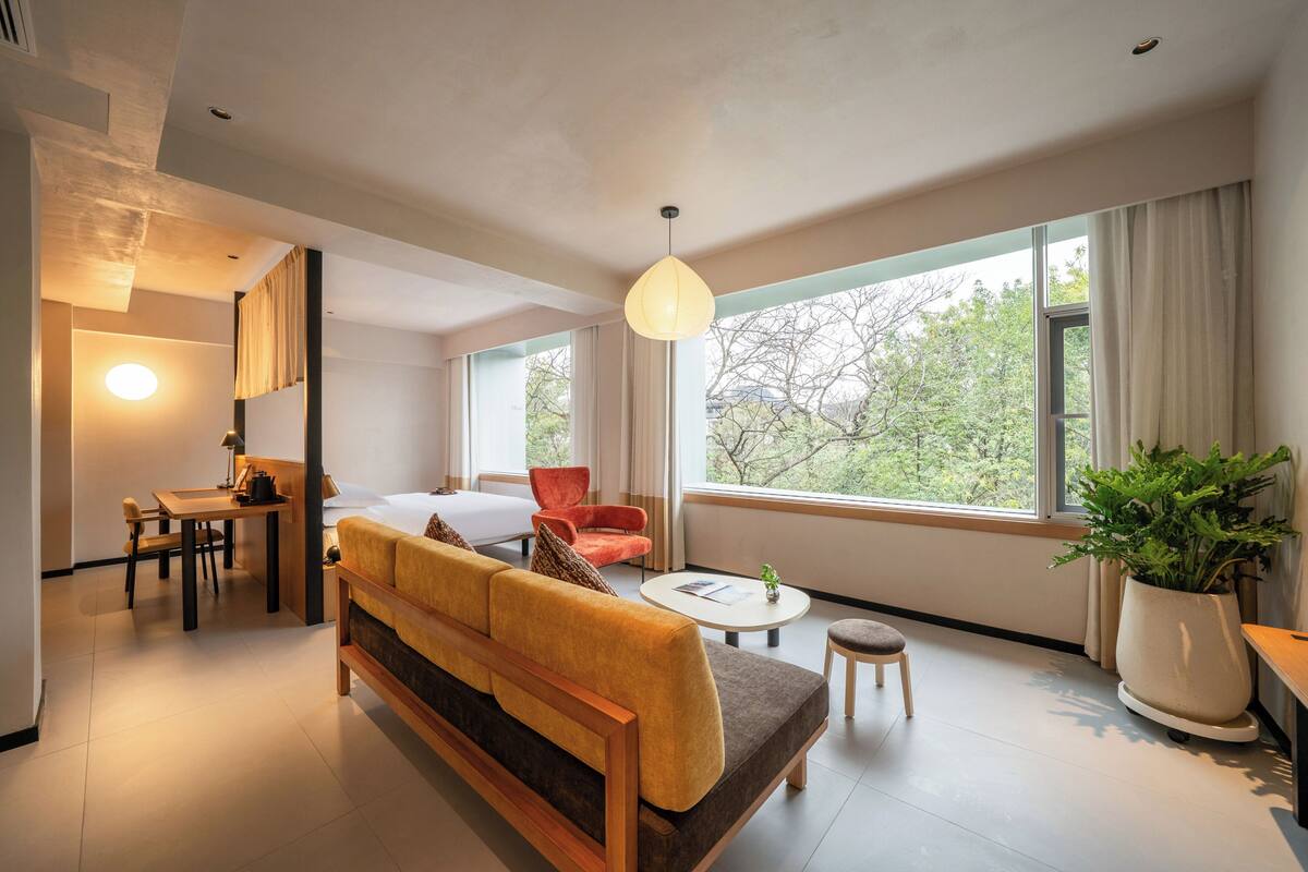 Una habitación de hotel moderna con sofá, mesa de comedor, sillas, un taburete pequeño, una planta en maceta y grandes ventanales con vistas a árboles.