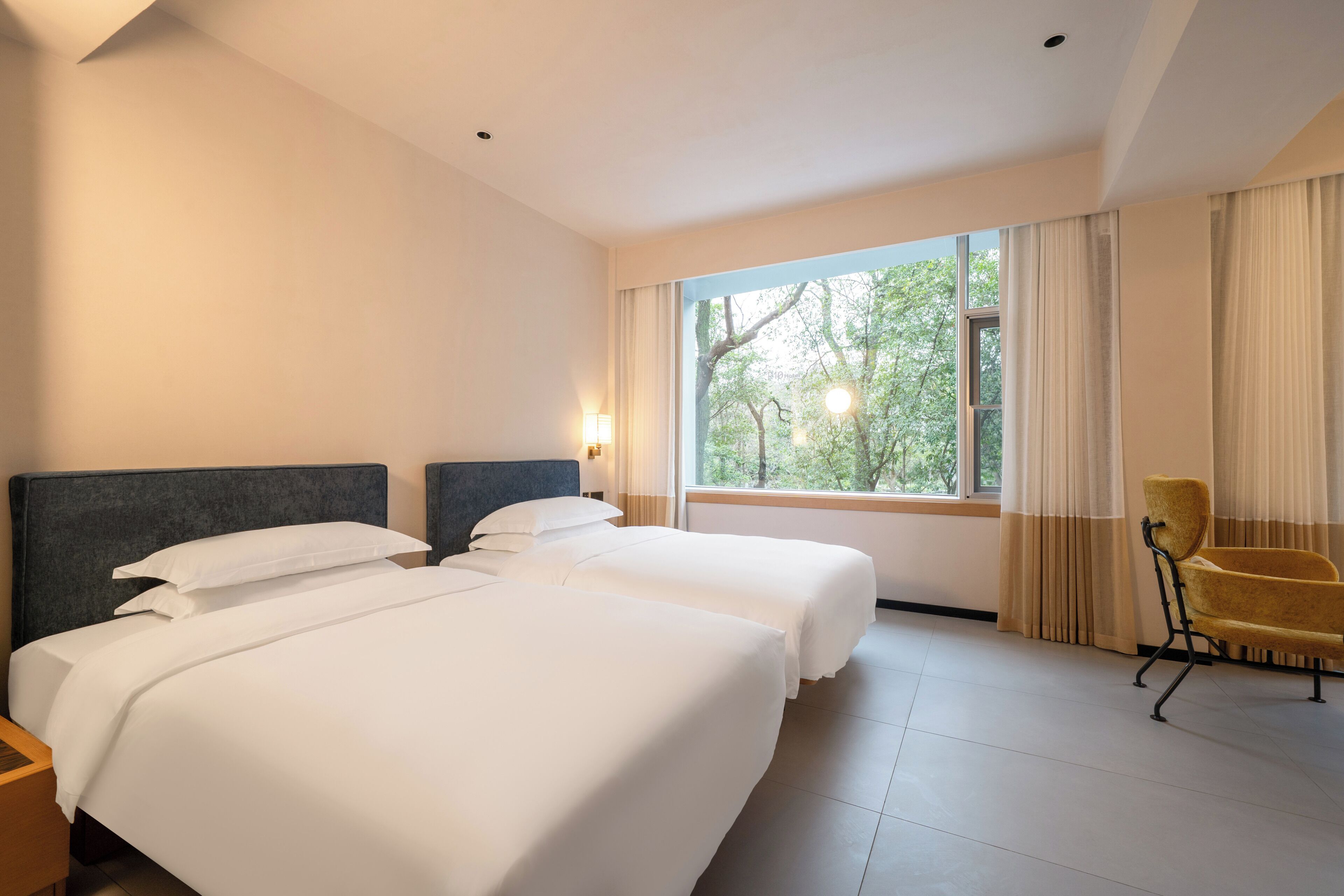 Camera d'albergo con due letti, una finestra vista alberi, una sedia e un comodino.