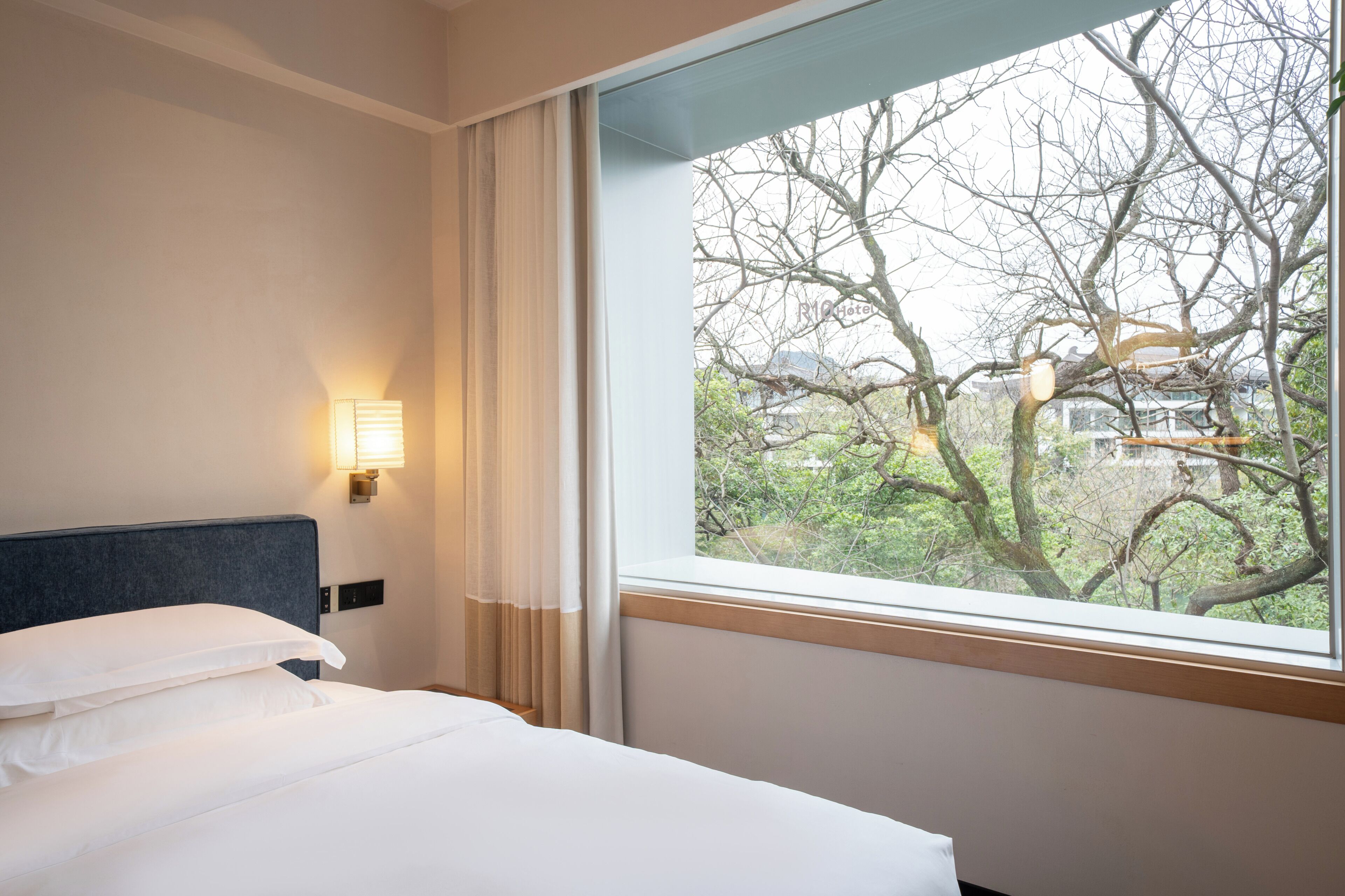 A hotel room with a bed, a window with a view of trees, and a wall-mounted light.