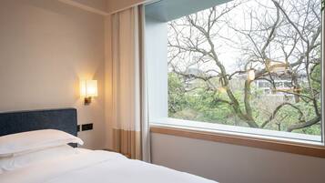 A hotel room with a bed, a window with a view of trees, and a wall-mounted light.