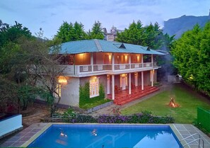 Una casa de dos pisos con piscina, rodeada de follaje y montañas al fondo.