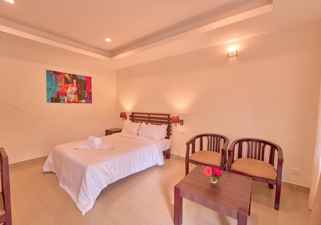 A hotel room with a bed, two chairs, a small table with a vase, and a painting on the wall.