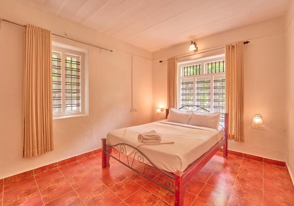 A single bed in a room with red tiled flooring, two windows with curtains, and a wall-mounted light fixture.
