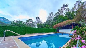 Una piscina con barandilla de metal, rodeada por un seto verde y una estructura con techo de paja.