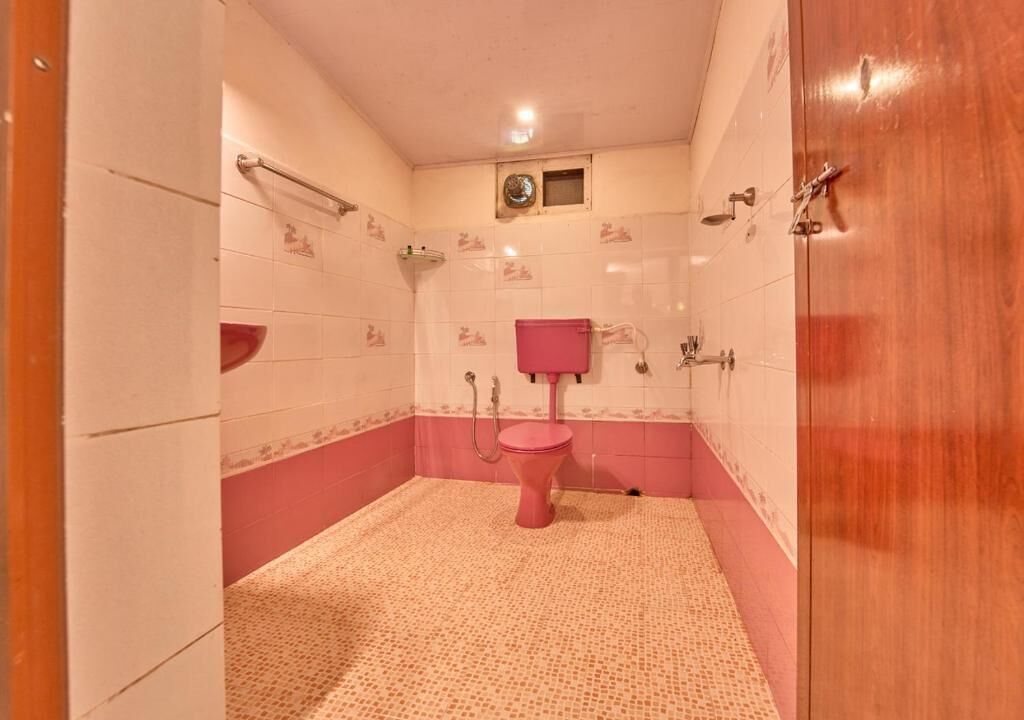 A bathroom with a pink toilet, white and pink tiled walls, a small window, and a wooden door.
