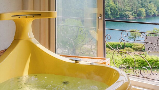 Una bañera de hidromasaje amarilla con vistas a un lago y vegetación a través de una ventana con barandilla de metal decorativa.