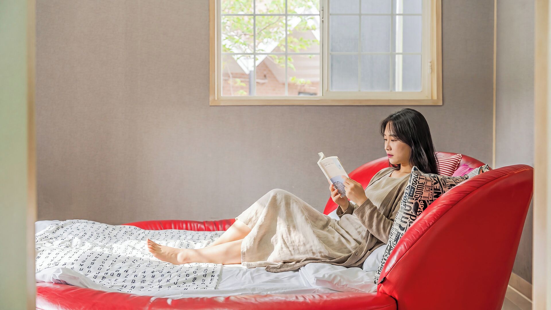 Une personne est assise sur un canapé rouge dans une pièce avec une fenêtre, et elle lit un livre.