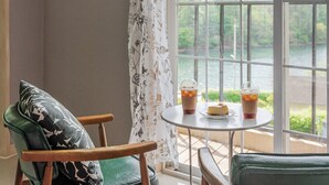 Una habitación con vista a un cuerpo de agua, una mesa con dos bebidas y una porción de pastel, y una silla con un cojín estampado.