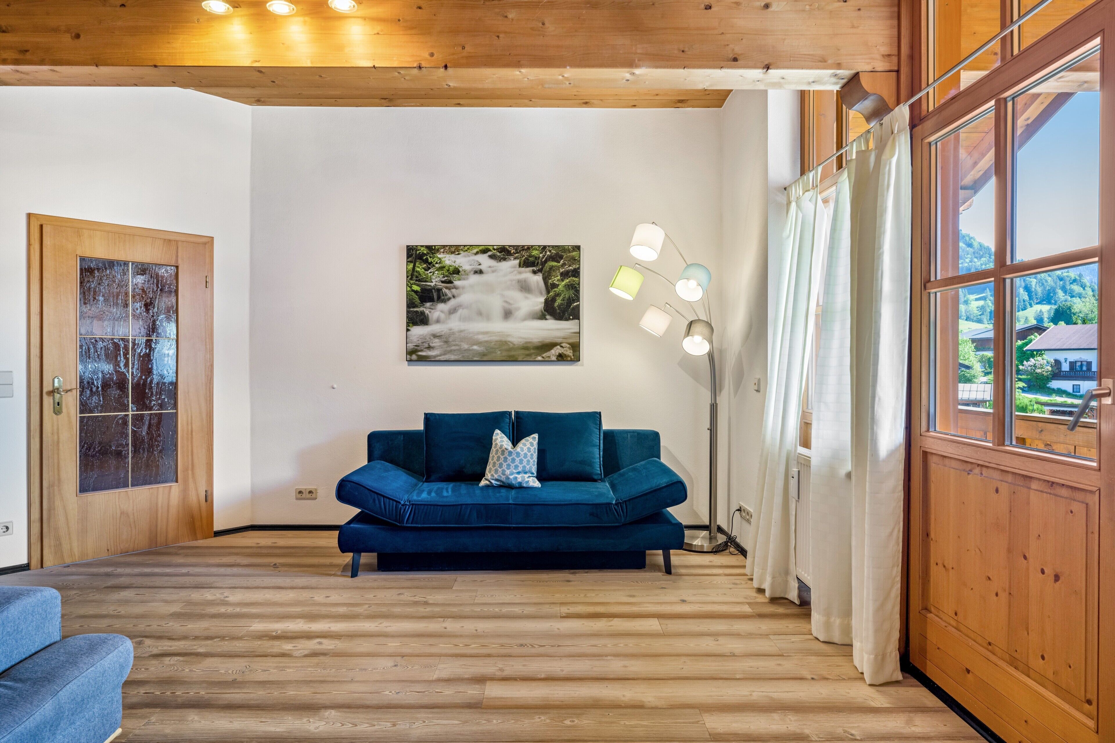 A room with a wooden floor, a blue sofa, a painting of a waterfall, a floor lamp, and a window with curtains.