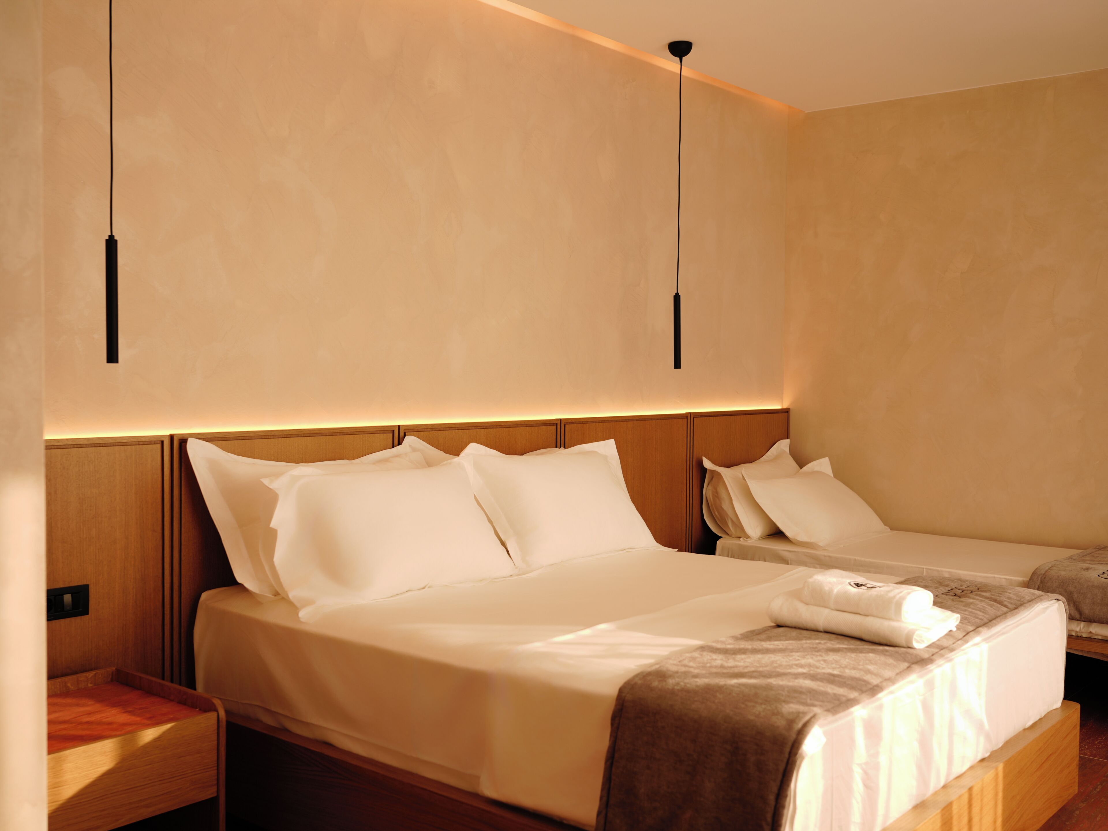 Quarto de hotel com uma cama grande, uma cama menor, cabeceira de madeira e iluminação pendente.