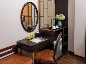 Habitación de hotel con un escritorio de madera, dos sillones tapizados en tono café, un espejo, una pequeña estantería con un jarrón de flores y un refrigerador.